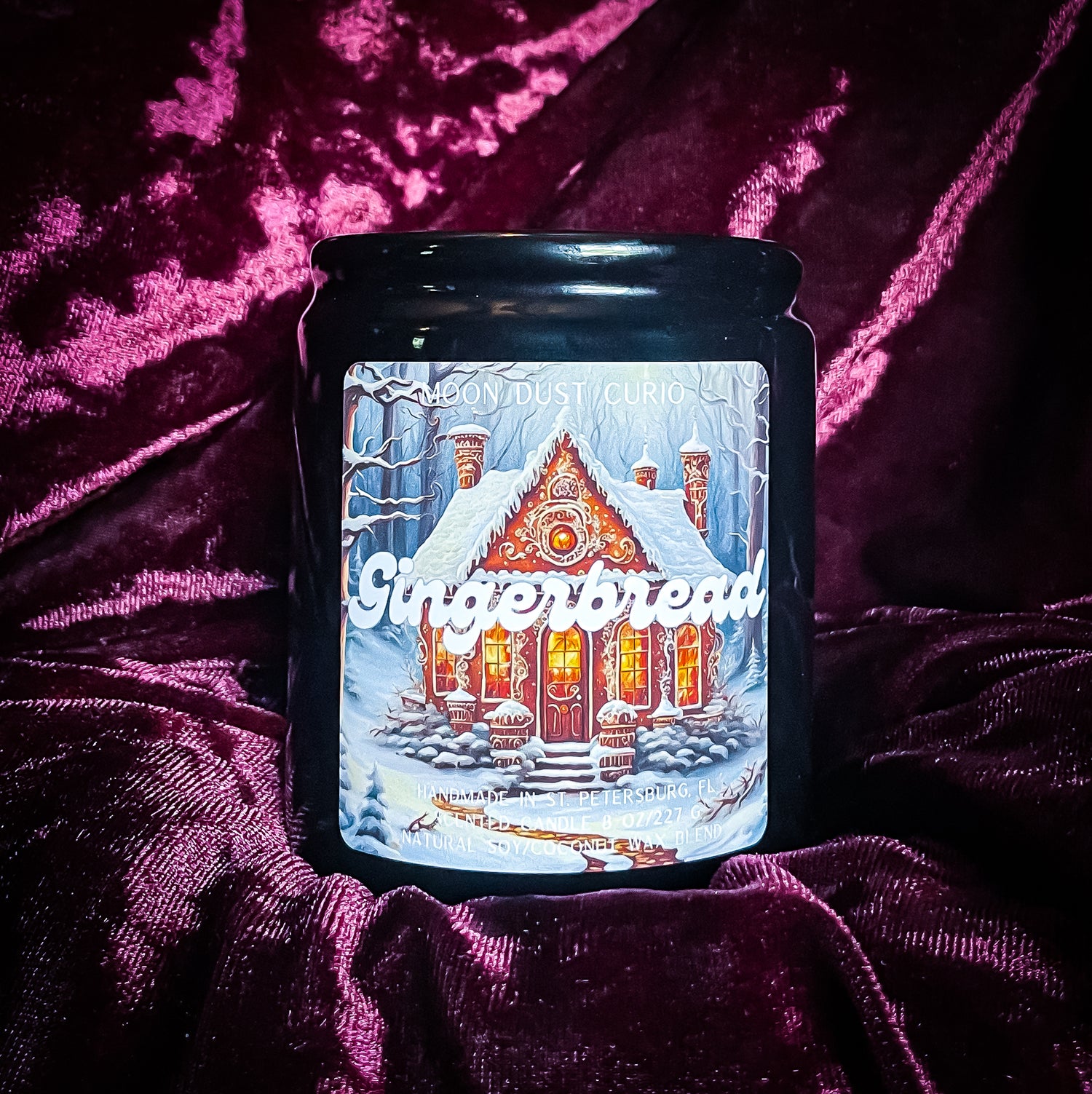 8 oz black ceramic candle with metal lid labeled “Gingerbread” in white script font; label features a snowy winter scene with a decorated gingerbread house surrounded by frosted trees.