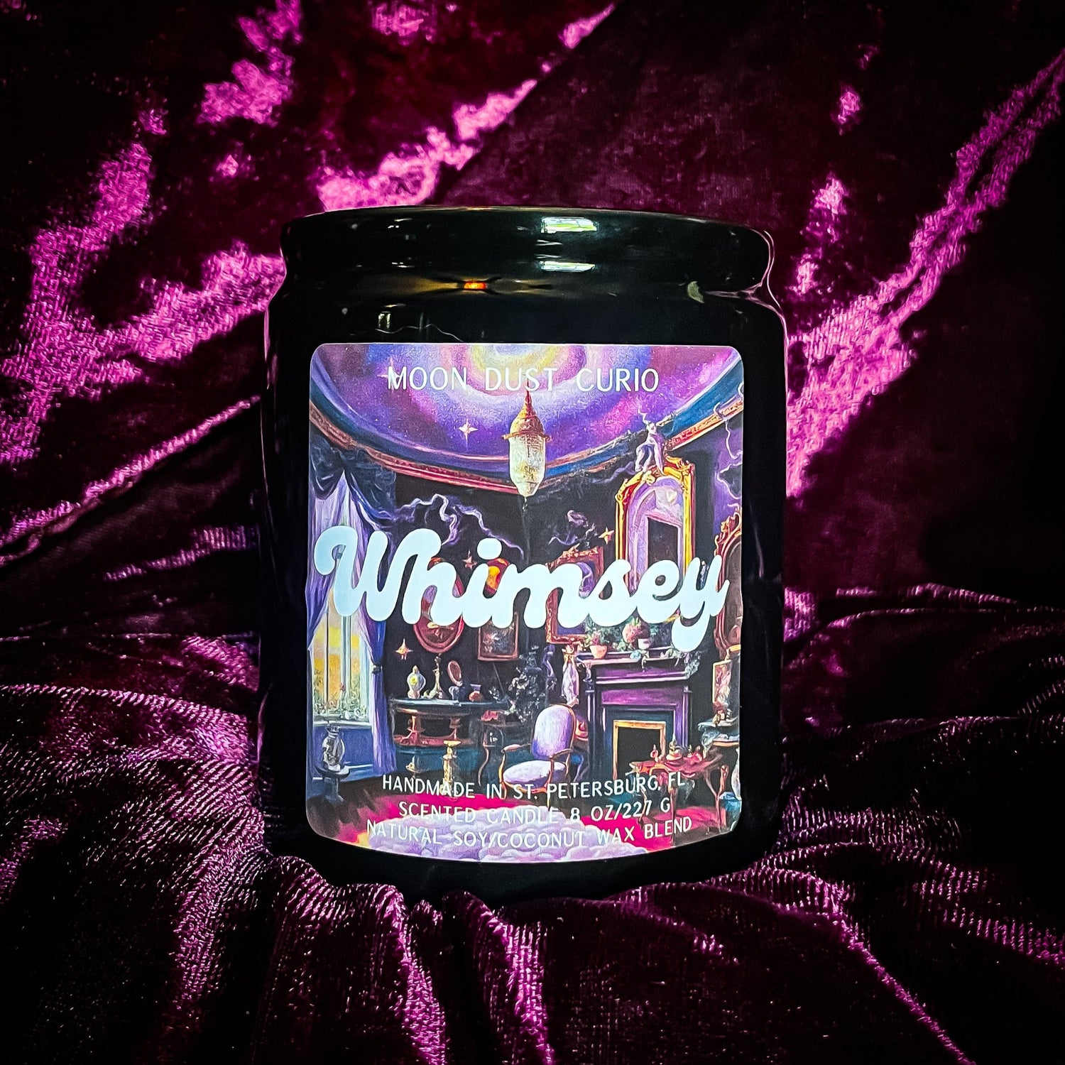 8 oz black ceramic candle with metal lid labeled “Whimsey” in playful script font; label features an ornate room with purple drapes, chandeliers, antique furniture, and shelves filled with curiosities.